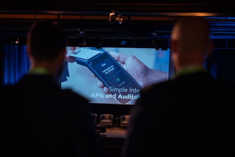 Dos asistentes a contraluz observan una pantalla de conferencia que muestra la validación de identidad ZAR ID en un dispositivo portátil