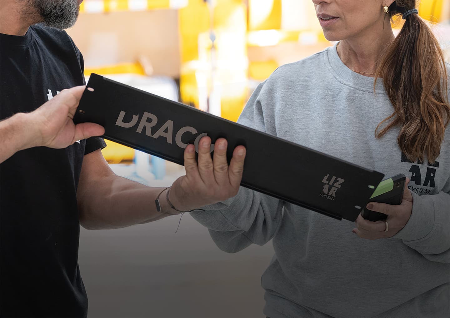 Zwei Personen in LIZAR-Systems-Kleidung reichen einander ein langes schwarzes Modul mit DRACO-Aufdruck in einem Produktionsbereich