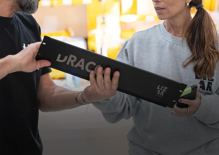 Dos personas con ropa de LIZAR Systems se pasan un módulo negro y alargado con la marca DRACO, en una zona de producción